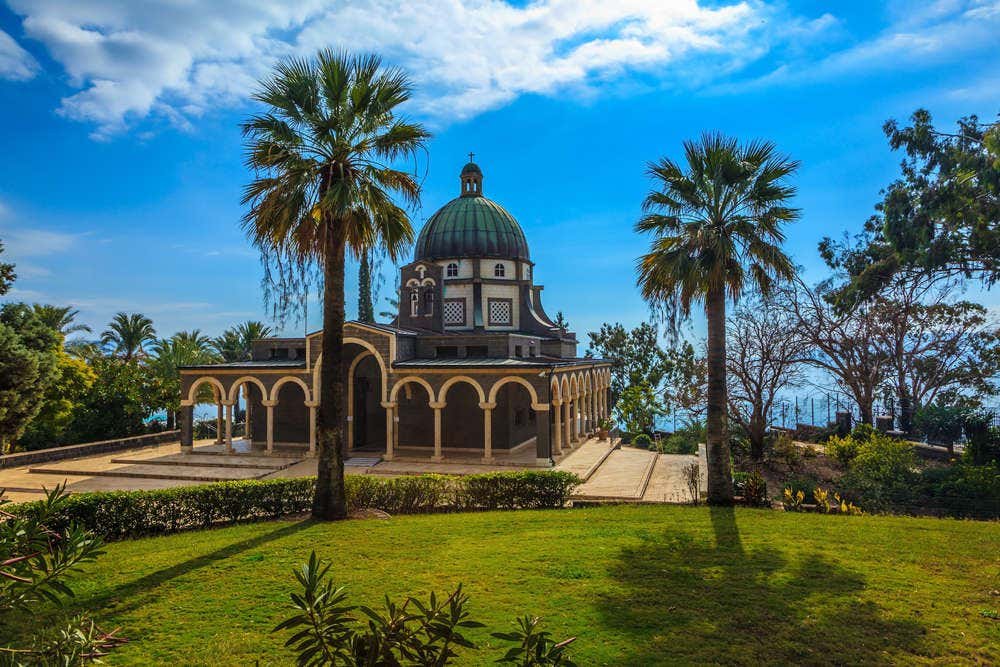Uma igreja histórica da Anunciação em Nazaré com teto abobadado é cercada por palmeiras e vegetação sob um céu azul parcialmente nublado.