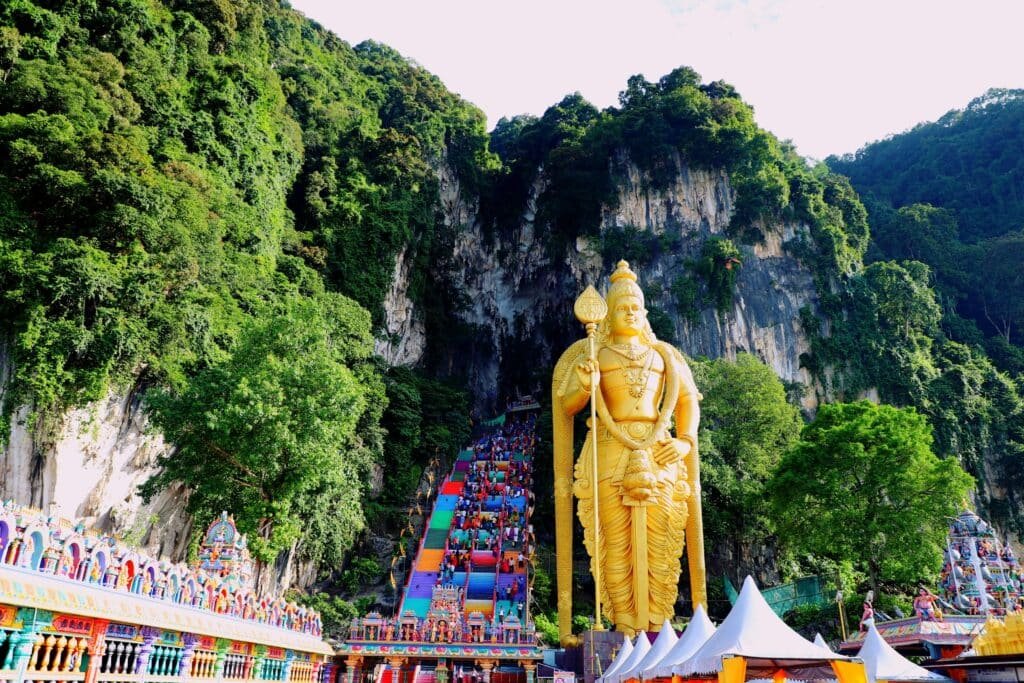 Templo hindu de  Batu Caves, Selangor na Indonésia com uma escultura dourada perto de uma rocha com escadas coloridas em meio a vegetação durante o dia. Representa chip celular Malásia.