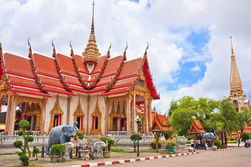 Um templo tailandês tradicional com telhados vermelhos ornamentados e torres douradas, cercado por estátuas de elefantes e árvores verdes sob um céu nublado.