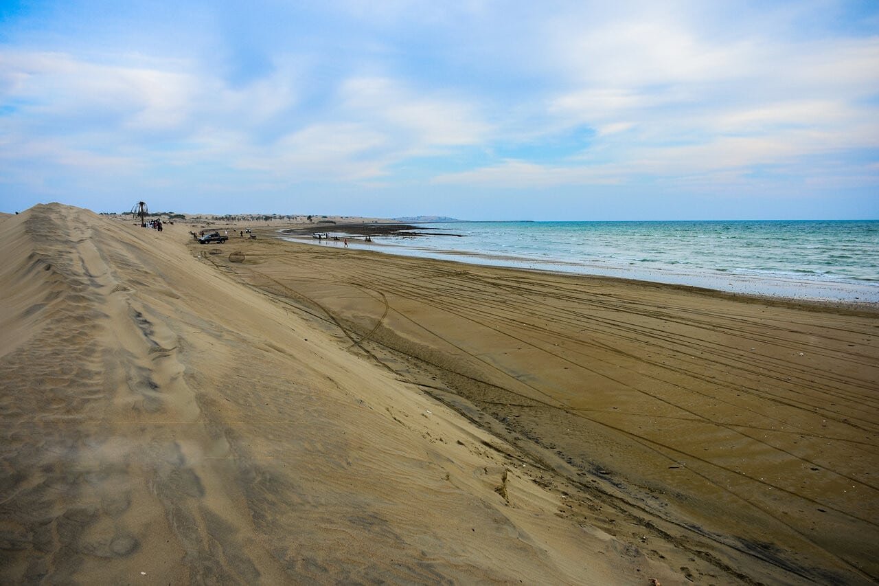Praia Darak Beach com marcas de pneus que levam ao mar sob um céu nublado. Estruturas distantes e pessoas visíveis à esquerda.