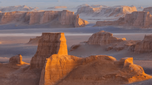 Formações de arenito em uma paisagem desértica no kaluts de Shahdad, com a suave luz do entardecer projetando sombras e destacando o terreno acidentado. Representa Irã.