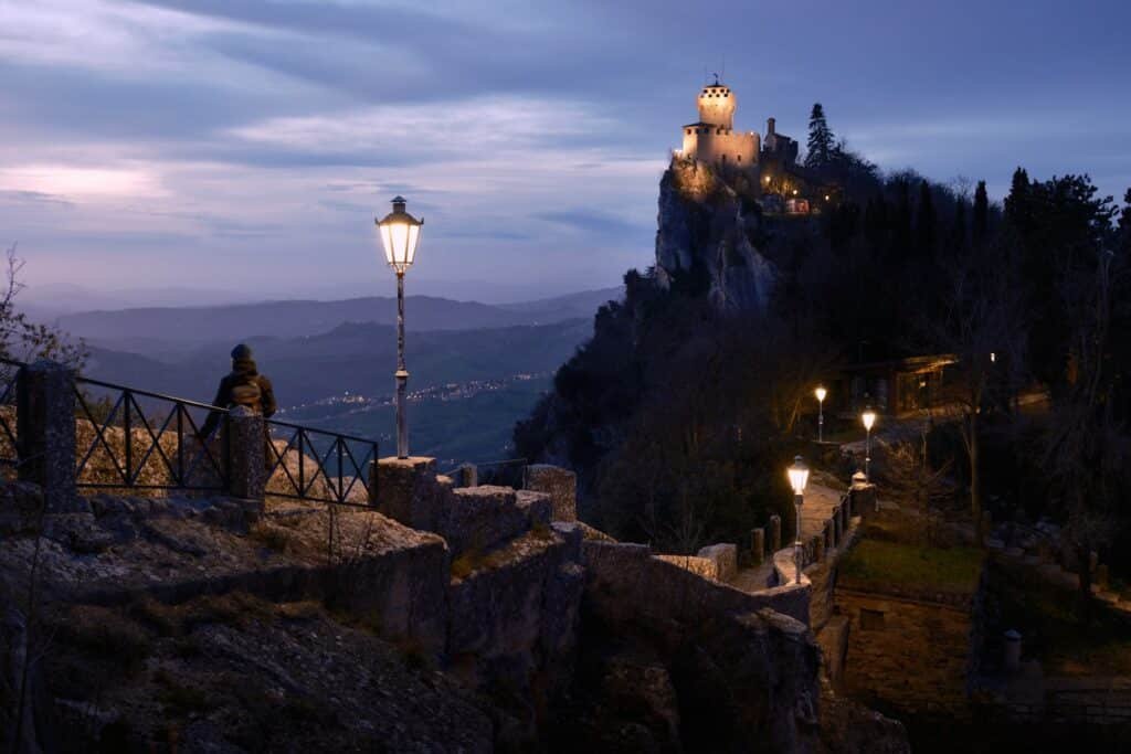 Um caminho de pedra leva a um castelo iluminado na encosta de um penhasco ao anoitecer, com postes de luz antigos e uma pessoa em pé perto de um corrimão. Ilustra o eSIM San Marino.