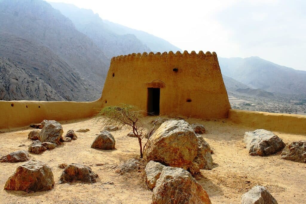Uma construção antiga em formato quadrado com uma porta, o local está no topo de uma montanha e há rochas na entrada, o local chama-se Forte Dhayah, para representar o que fazer em Ras Al Khaimah