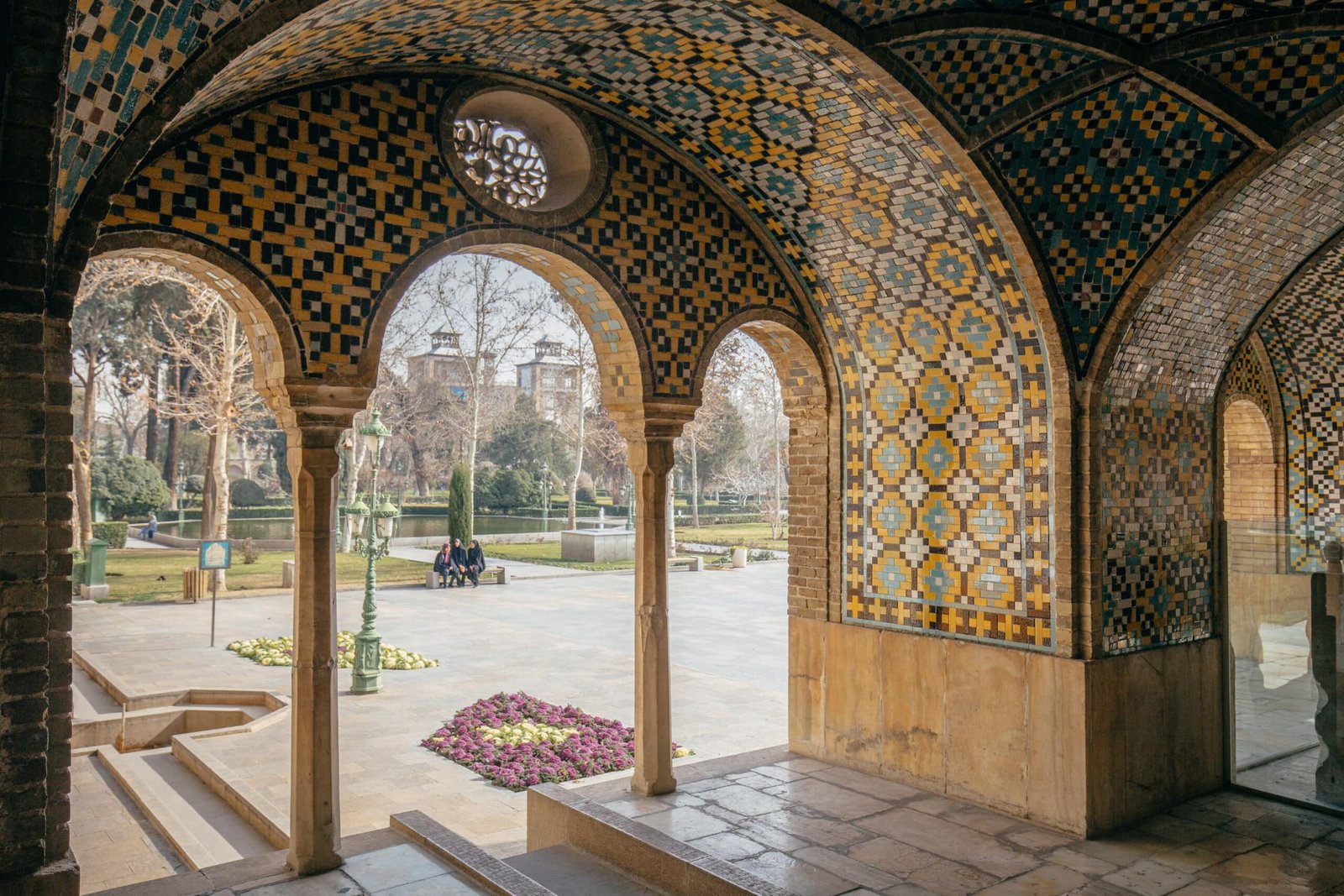 Arcos ornamentados com padrões de azulejos coloridos dão para um jardim com árvores e flores em um dia ensolarado no Golestan Palace.
