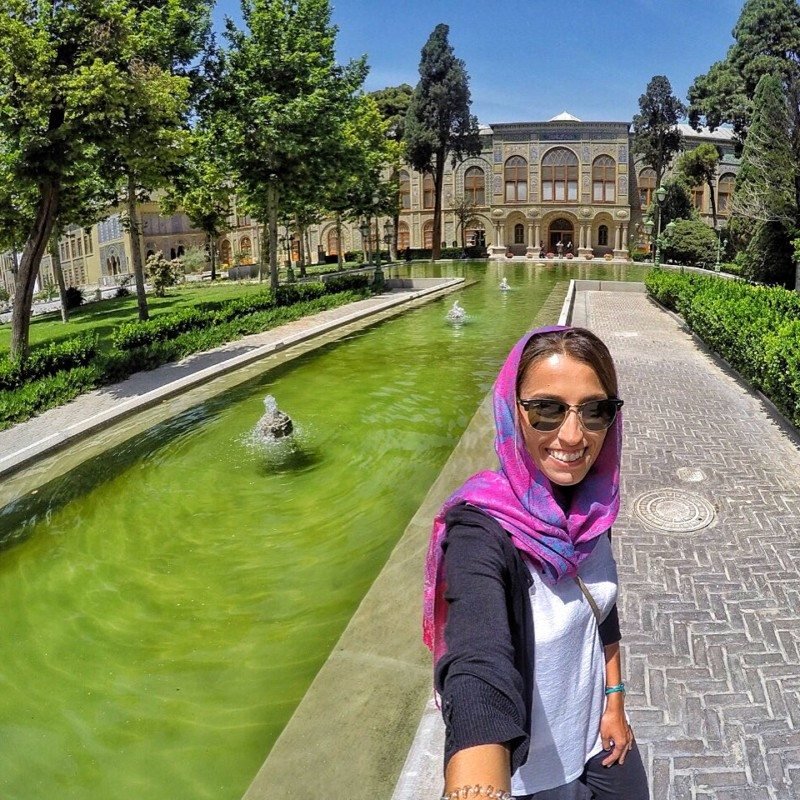 Uma pessoa tira uma selfie em frente ao Golestan Palace com uma longa piscina verde e árvores em um dia ensolarado. Imagem para ilustrar post sobre o que fazer no Irã.