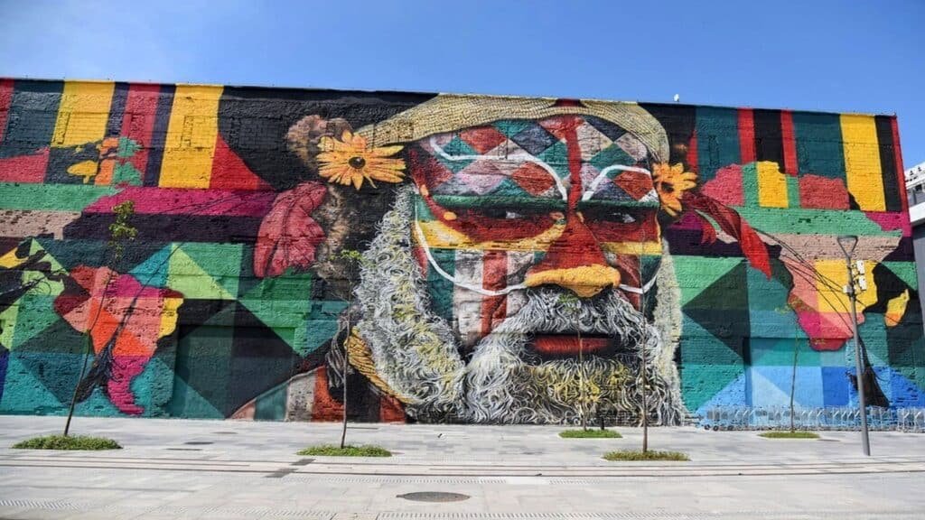 Grande mural do rosto de um homem barbudo com padrões geométricos coloridos e flores, pintado na lateral de um prédio de Hilus. Imagem para ilustrar post sobre eSIM Paupua Nova Guiné.