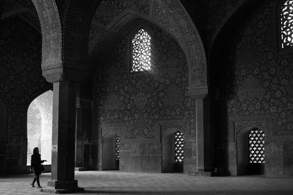 Foto em preto e branco do interior de uma mesquita grande no Irã e ornamentada, com uma pessoa de pé perto de uma coluna, iluminada por janelas estampadas.