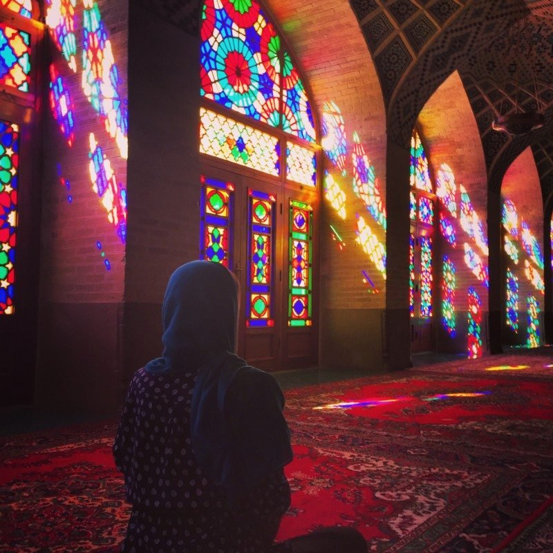 Uma de nossas colunistas com um lenço azul na cabeça está sentada em um tapete estampado em uma sala com vitrais vibrantes projetando padrões de luz coloridos na Mesquita Nasir ol Molk. Imagem para ilustrar post sobre o que fazer no Irã.