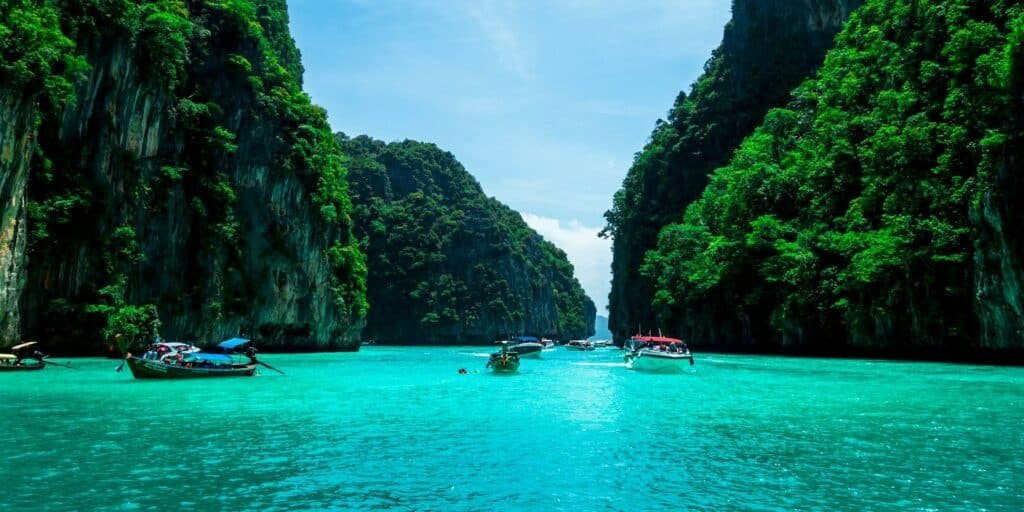 A imagem mostra um cenário de águas cristalinas em tons de azul-turquesa, cercadas por altos penhascos cobertos de vegetação densa e verdejante. No centro, várias pequenas embarcações, incluindo barcos típicos tailandeses, estão ancoradas ou navegando pela água. O céu está claro e azul, com poucas nuvens visíveis. Esta paisagem é típica das áreas costeiras de Phuket, na Tailândia, onde a natureza exuberante encontra o mar tranquilo.