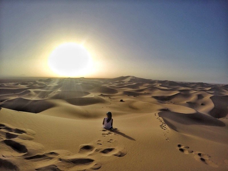 Uma pessoa está sentada em uma duna de areia no Mobarakeh Bafgh, com o sol baixo no céu projetando longas sombras na areia.