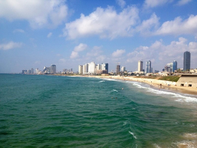 Praia em Tel Aviv no primeiro plano da imagem, mostrando uma faixa branca e areia ao fundo e a cidade com vários prédios atrás. Ilustra o post sobre o que fazer em Israel.