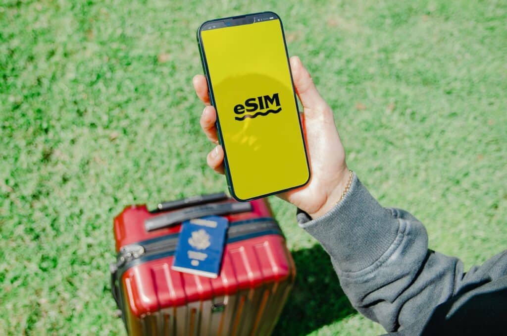 Uma pessoa segura um smartphone com a inscrição "eSIM" em uma tela amarela, acima de uma mala vermelha e um passaporte, na grama verde. A foto é parte do post de eSIM Abu Dhabi.