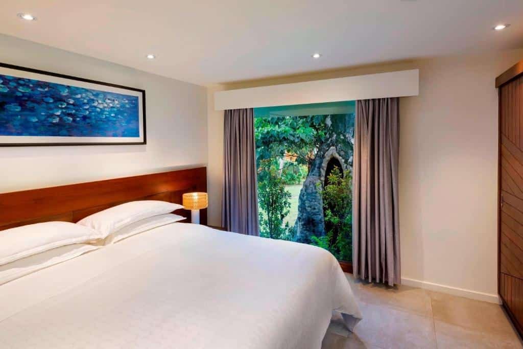 Quarto do Sheraton Resort & Spa, Tokoriki Island  com cama de casal, cômoda de madeira do lado esquerdo com luminária e janelas amplas com cortinas cinzas com vista para o jardim.