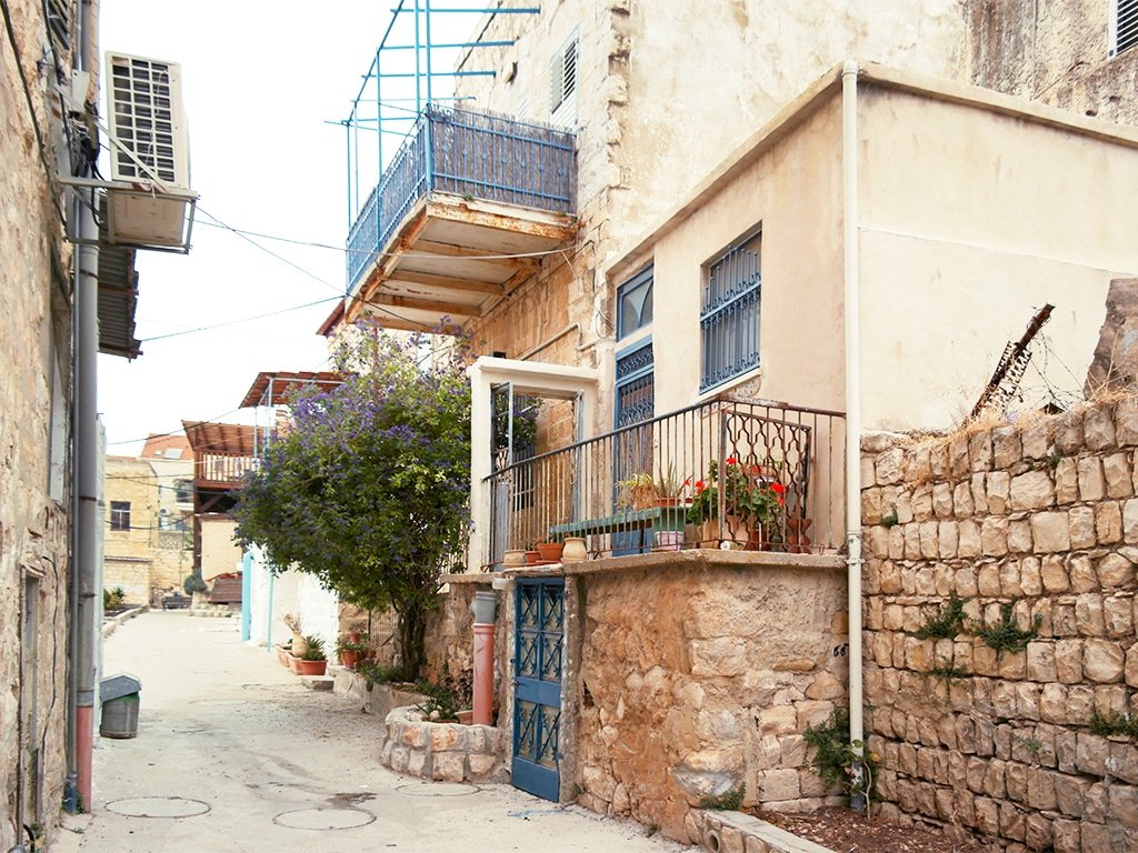 Beco estreito em Safed (Israel) com prédios de pedra, sacadas de metal azul e plantas em vasos em saliências. Unidade de ar condicionado visível à esquerda.