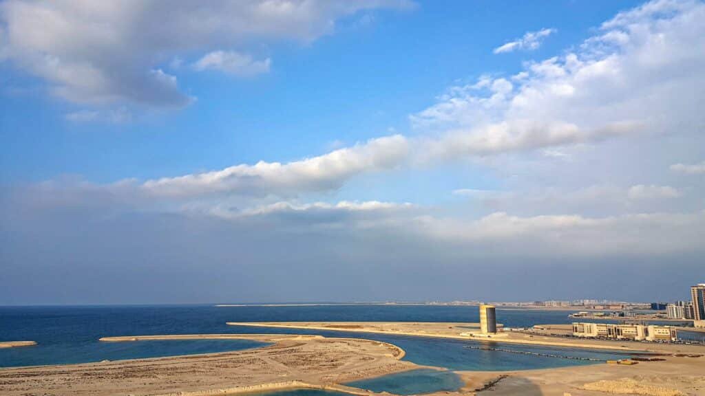 Céu azul com nuvens brancas, sob um mar azul cristalino, com uma faixa de terra com algumas construções de prédios. 