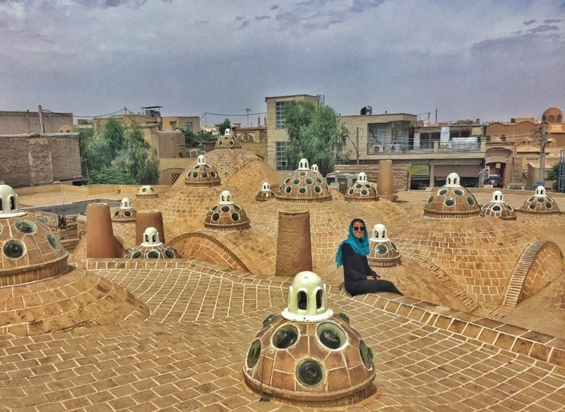Uma pessoa sentada em um telhado de tijolos tradicional com cúpula e aberturas circulares de vidro, cercada por um céu nublado e edifícios no Sultan Amir Ahmad Bathhouse.