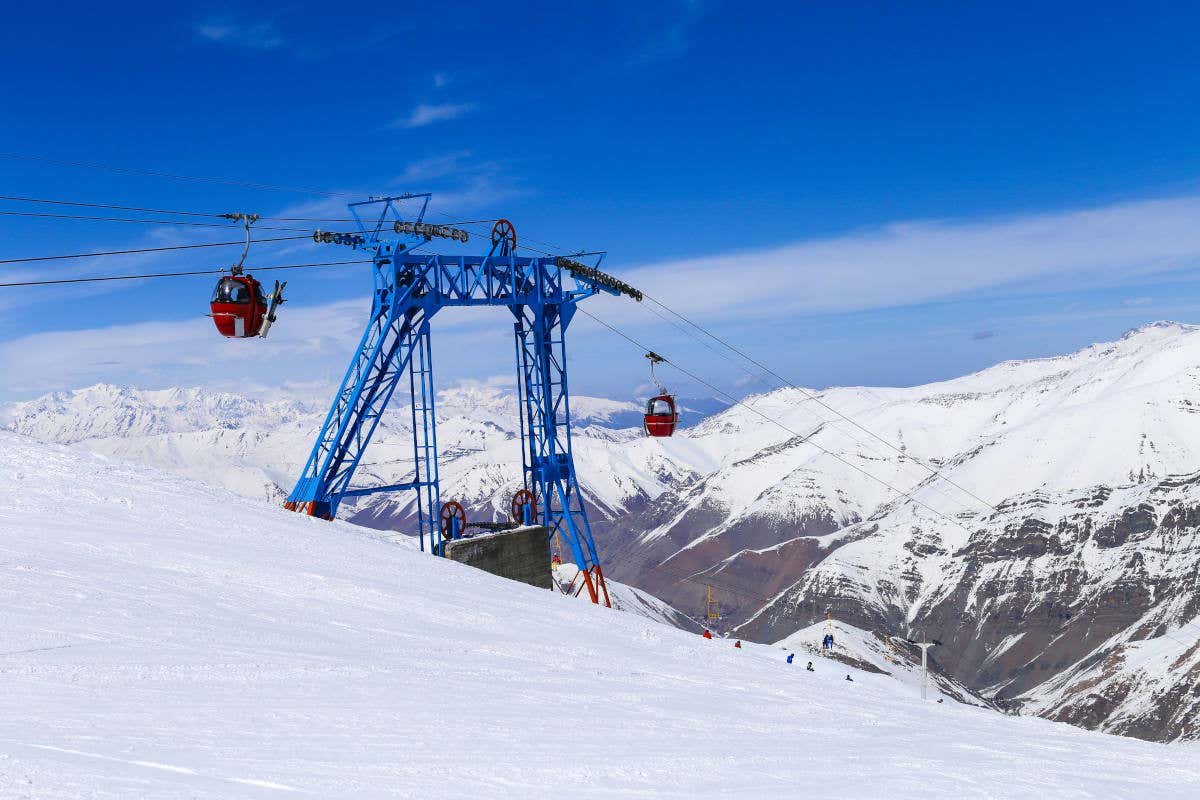 Uma estrutura de teleférico azul no Dizin International Ski Resort nevada com gôndolas vermelhas, tendo como pano de fundo picos cobertos de neve e um céu azul. Imagem para ilustrar post sobre o que fazer no Irã.