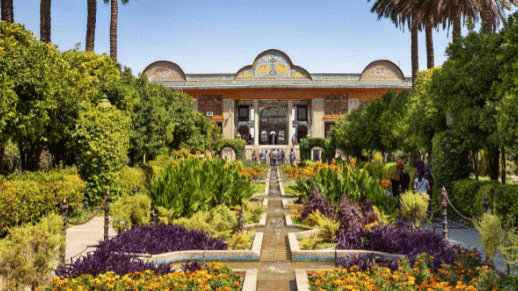 Um jardim persa histórico com flores vibrantes em Shiraz, fontes simétricas e um edifício tradicional sob um céu azul claro.