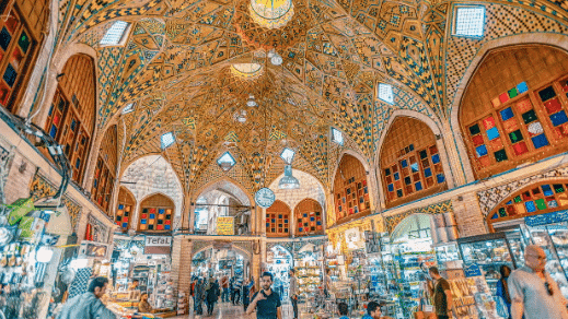 Pessoas caminhando e visitando lojas dentro de um bazar vibrante e ornamentado com tetos abobadados e azulejos coloridos no Teerã. Representa Irã.
