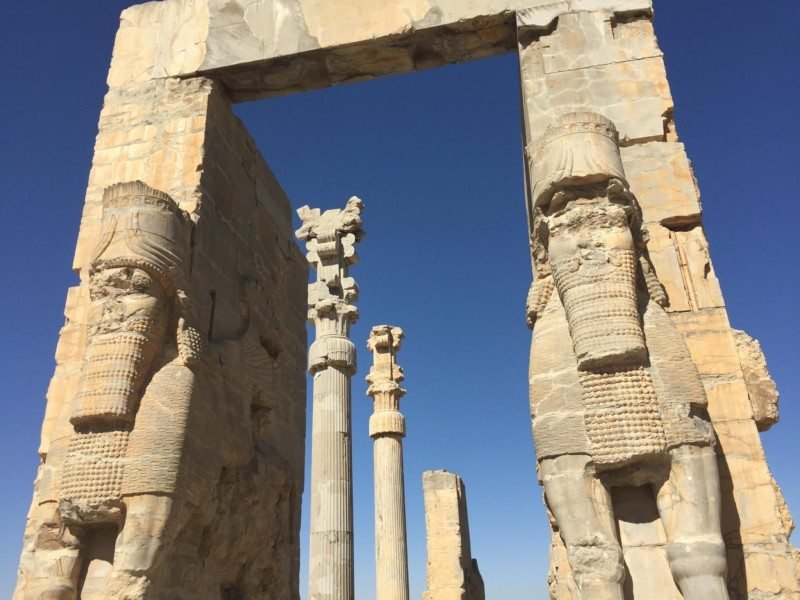 Imagem de ruínas persas durante o dia no Irã, com colunas de concretos em cada lado da imagem com imagem de figuras. Representa Irã.