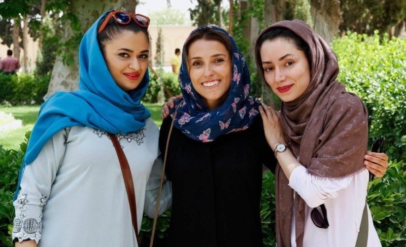 Imagem de três mulheres a frente com lenço na cabeça, durante o dia em frente a um jardim. Representa Irã.