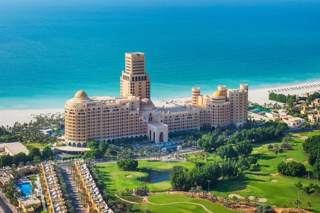 Prédio do Waldorf Astoria Ras Al Khaimah com vista para um mar muito azul e de areia branca, aos fundos da propriedade, há um amplo gramado com árvores e um pequeno lago